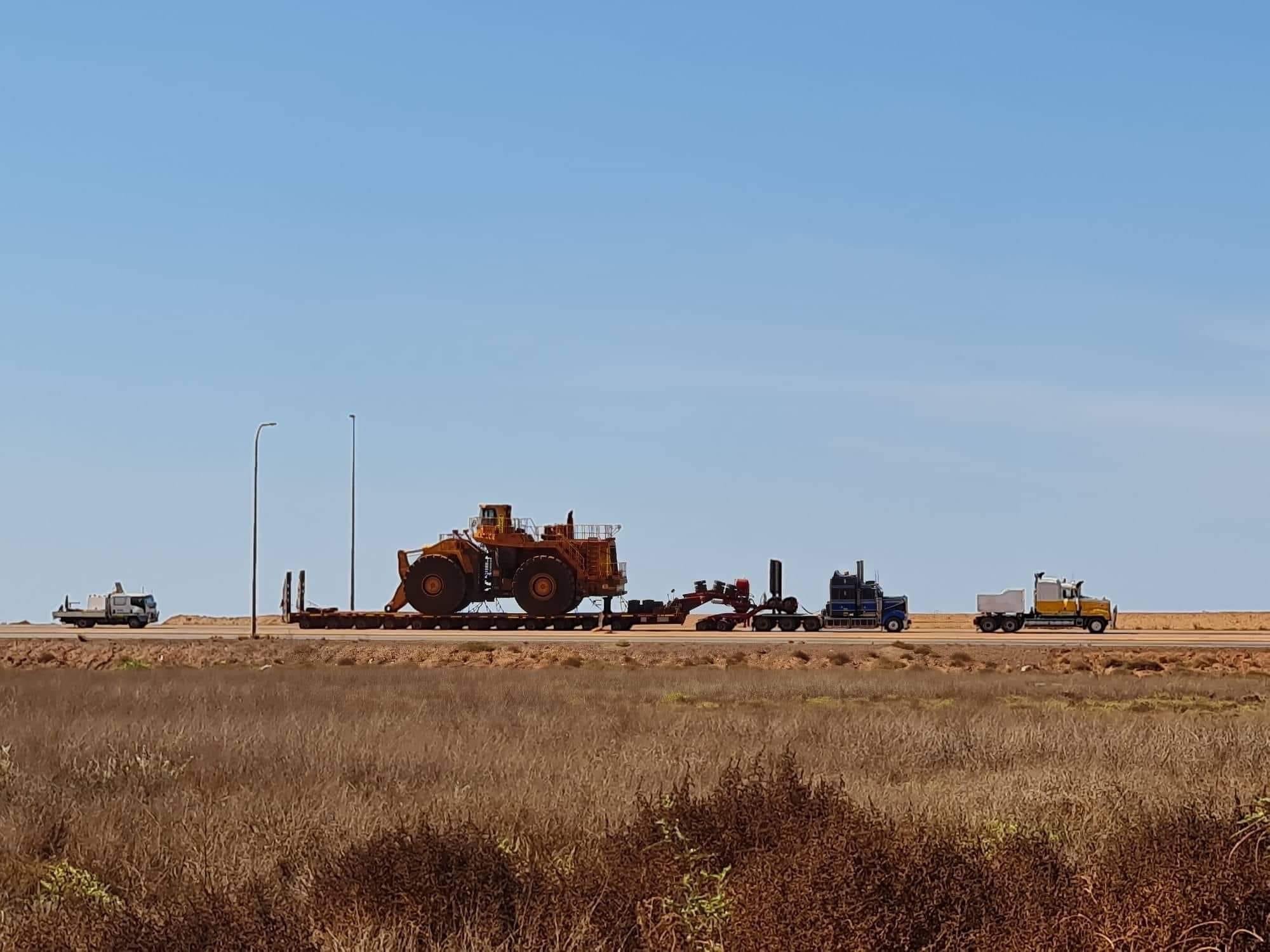 Side view of a heavy vehicle convoy featuring three trucks, one with a long trailer towing a earthmoving machine, moving along a road