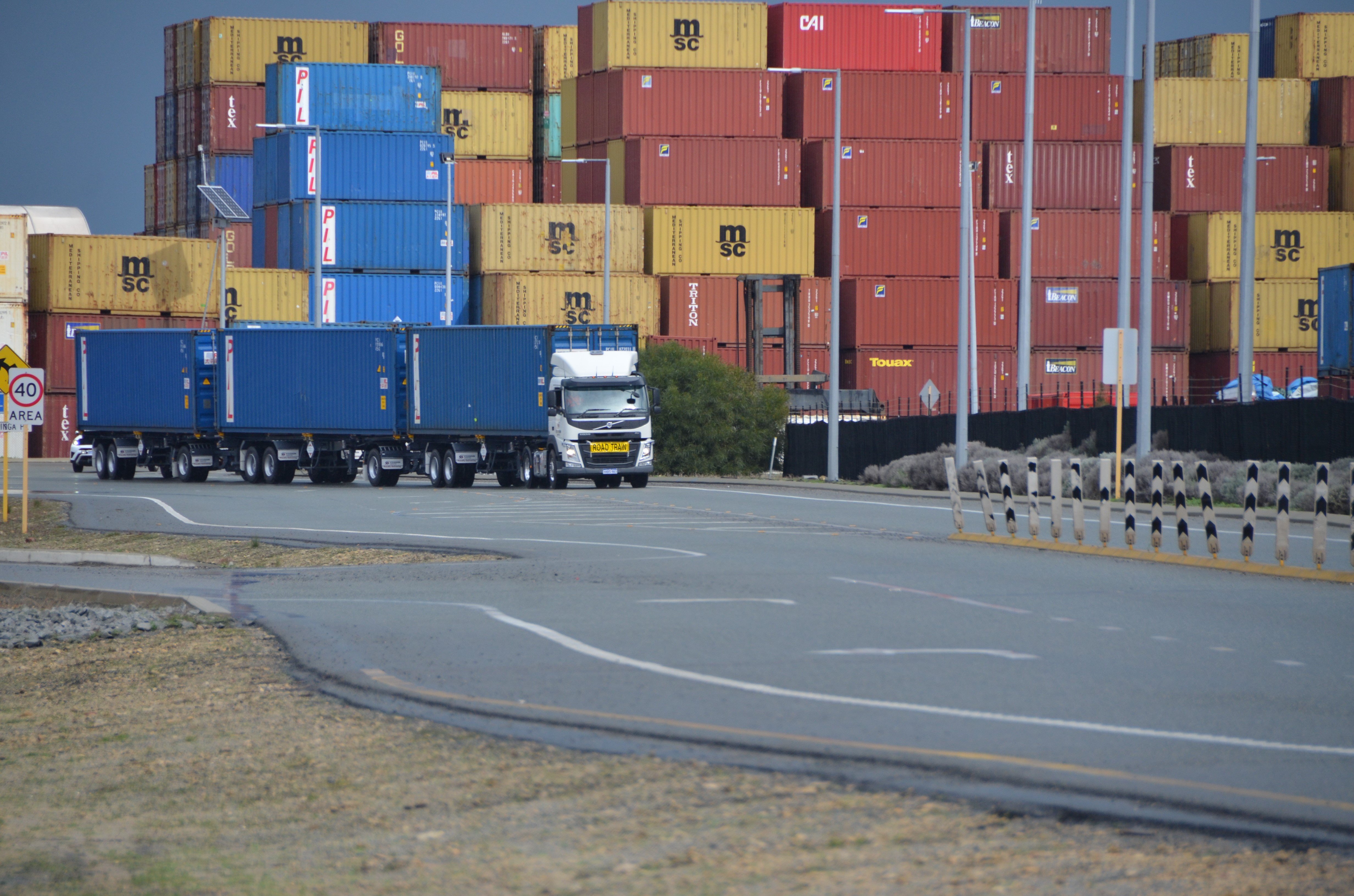 Blue road train towing three blue containers driving along a road in front of a shipping port with a stack of sea containers