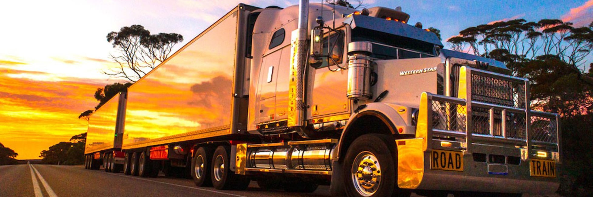 Long white truck on a road with the sunset in the background and reflecting off the side of the truck