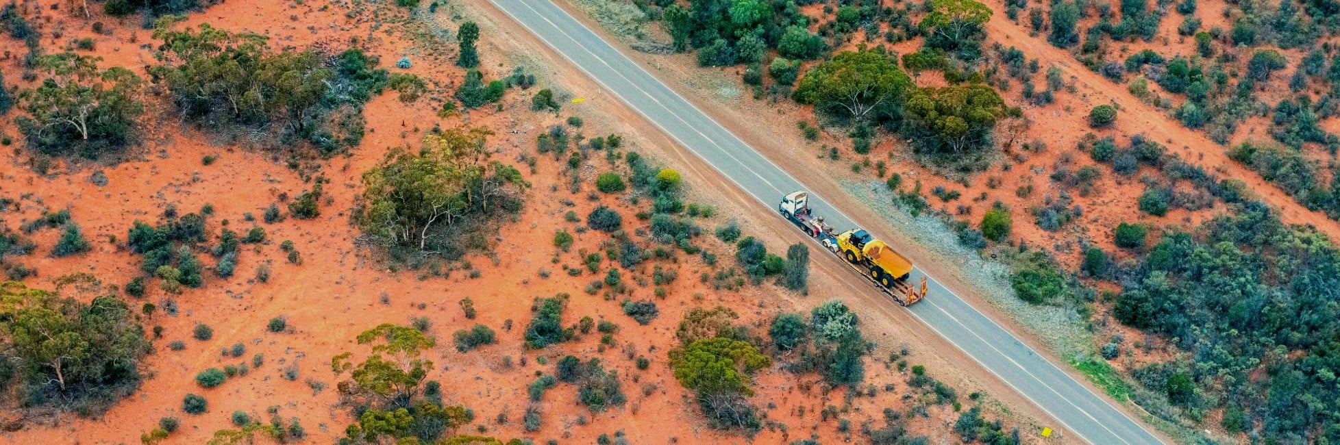 Long Truck with a tipper truck on it's trailer driving along a road with red dirt and shrubs in the background